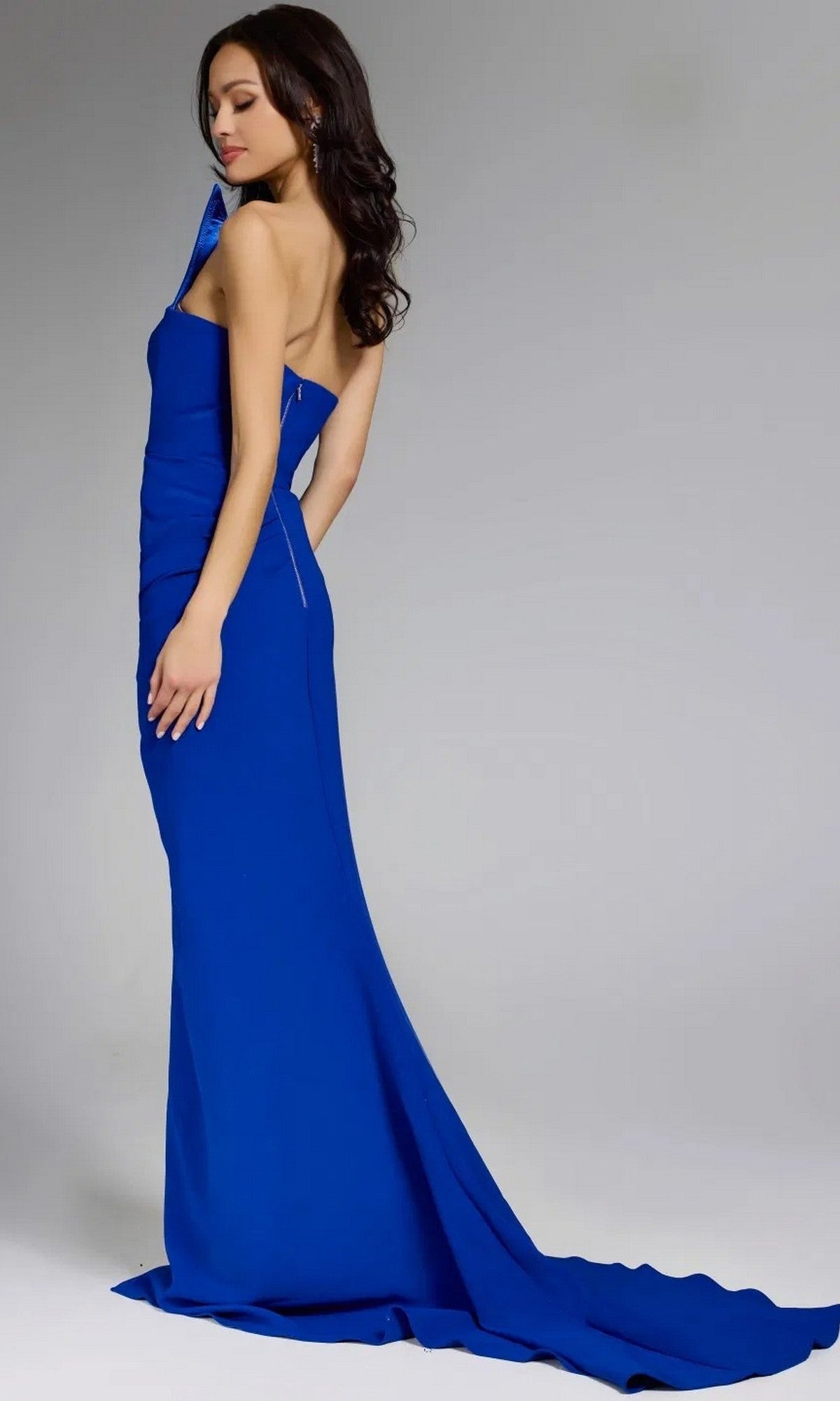 Model wearing the Jovani 44001 long prom dress, a royal blue floor-length gown with an asymmetrical neckline and long train, standing sideways against a plain light gray background.